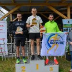 DSC_6590 Na podium stoją trzej mężczyźni z nagrodami. Tło stanowi drewniana konstrukcja, obok banery i zieleń. Dwaj trzymają flagę z napisem MIĘSKI KLUB BIEGACZA DREPTAK.