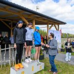 DSC_6594 Na podium stoją trzy kobiety, z których jedna otrzymuje nagrodę. Kobiety ubrane są w sportowe stroje, dominują kolory czarny i niebieski. W tle widać zadaszoną scenę i grupę obserwujących osób.