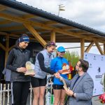 DSC_6598 Na podium trzy kobiety otrzymują trofea. Mają sportowe stroje, jedna w niebieskiej czapce. W tle drewniana konstrukcja i baner.