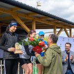 DSC_6601 Na podium stoją trzy kobiety, jedna trzyma puchar, druga kwiaty. Tło to drewniana konstrukcja i błękitne niebo. Dookoła są klaskający widzowie.