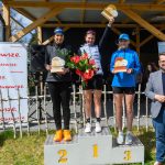 DSC_6607 Na podium stoją trzy kobiety z trofeami, uśmiechają się. Tło tworzy drewniana altanka i baner „Mazowsze”. Wiosenna sceneria z kwiatami.