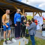 DSC_6613 Na podium stoją trzy osoby w sportowych strojach podczas ceremonii wręczenia nagród. W tle drewniana wiata i biały baner na łące. Mężczyzna wręcza kobiecie trofeum, obok stoją dwie młode osoby z medalami.