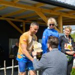 DSC_6616 Na zdjęciu mężczyzna w sportowym stroju odbiera trofeum. W tle drewniana konstrukcja, słońce i zielone drzewa. Obok stoją dwie kobiety oraz kilka osób w tle.