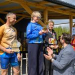 DSC_6619 Na zdjęciu są osoby stojące na podium i odbierające nagrody. Tło to drewniana altana i zielone drzewa. Osoby mają medale i statuetki.