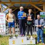 DSC_6623 Trzy osoby na podium, trzymają puchary. Tło z drewnianą konstrukcją i plakatem Mazowsze. Słoneczny dzień, trawa i niebo.