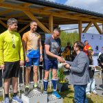 DSC_6625 Trzech mężczyzn na podium, w sportowych strojach, z medalami. Tło to drewniana konstrukcja i roślinność. Osoba wręczająca nagrody, w okularach i marynarce.