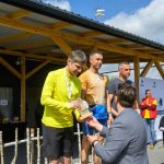 DSC_6627 Zawodnicy na podium otrzymują nagrody na zewnątrz. Mężczyzna w żółtej bluzie przyjmuje trofeum. W tle konstrukcja z drewna i niebo z chmurami.