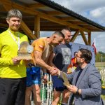 DSC_6628 Na zdjęciu czterech mężczyzn; jeden wręcza trofea. W tle drewniana wiata i niebo. Przewaga jasnych kolorów.