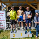 DSC_6633 Trzech mężczyzn na podium z trofeami; jeden w żółtej koszulce, drugi w beżowo-niebieskiej, trzeci w granatowej. Tło z banerami na zewnątrz, jasnozielona trawa. Stoją obok nich mężczyźni w maryn