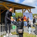 DSC_6637 Na zdjęciu mężczyzna wręcza nagrodę kobiecie na zewnątrz drewnianego budynku. Tło jest zielone z drewnianym płotkiem, kilka osób w tle. Atmosfera jest słoneczna i radosna.