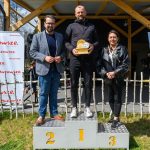 DSC_6640 Troje ludzi stoi na podium: miejsca 1, 2, 3. Ubrani w ciemne kurtki, otoczeni drewnianym ogrodzeniem, z banerem Mazowsze w tle.