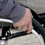 Ręka na kole wózka inwalidzkiego. Szare ubranie, metalowy obręcz koła, tło neutralne. Skupienie na detalu i wsparciu.