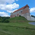 0Z0vvPDf Ceglany zamek z czerwoną dachówką stoi na zielonym wzgórzu. W tle niebo z białymi chmurami. Obok prowadzi wąska droga.