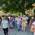 A1oCFr0J Grupa osób spaceruje po brukowanej ulicy, częściowo zacieniona przez drzewa. W tle widać budynki i zadaszenia. Ubrania w jasnych i ciemnych kolorach.