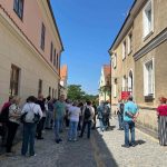CT__iYZ Grupa ludzi stoi na wąskiej, brukowanej uliczce między pastelowymi budynkami. Tło to czyste, niebieskie niebo. Po prawej stronie widoczne są białe okna i czerwony szyld.
