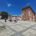 WT7trdZ1 Zdjęcie przedstawia brukowany rynek z ceglanym budynkiem po prawej stronie. Niebieskie niebo i kilka białych chmur w tle. Ludzie spacerują po placu otoczonym kamienicami.