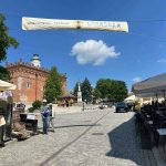 YALnSETR Brukowany rynek z czerwonym gotyckim budynkiem w tle, błękitne niebo z białą chmurką. Banner na festiwal winny, stoliki po prawej.